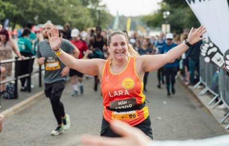 Woman in a Place2be running vest, taking part in a race and running past with her arms in the air.