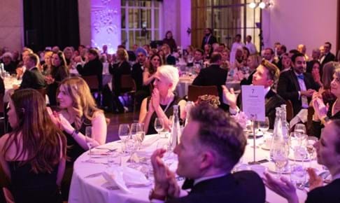 Guests at Place2Be's Arts and Minds Gala. They are sitting on round tables in a formal setting, and are applauding.