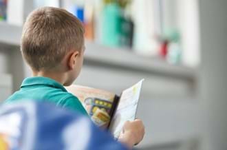 A young boy sitting down in a classroom, facing away from the camera. He is reading a book.