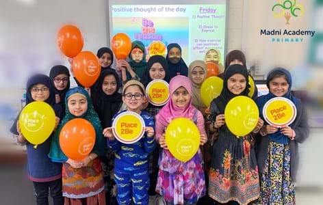A class of primary school pupils holding yellow and orange Place2Be balloons. They are standing in at the front of their classroom.