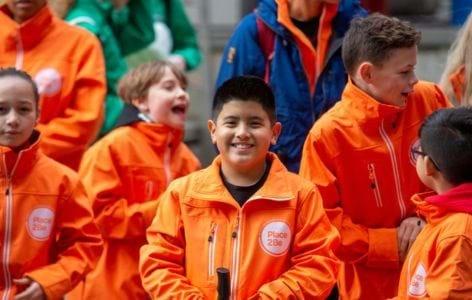 Group of diverse children smiling and wearing Place2Be orange jackets at a fundraising event.