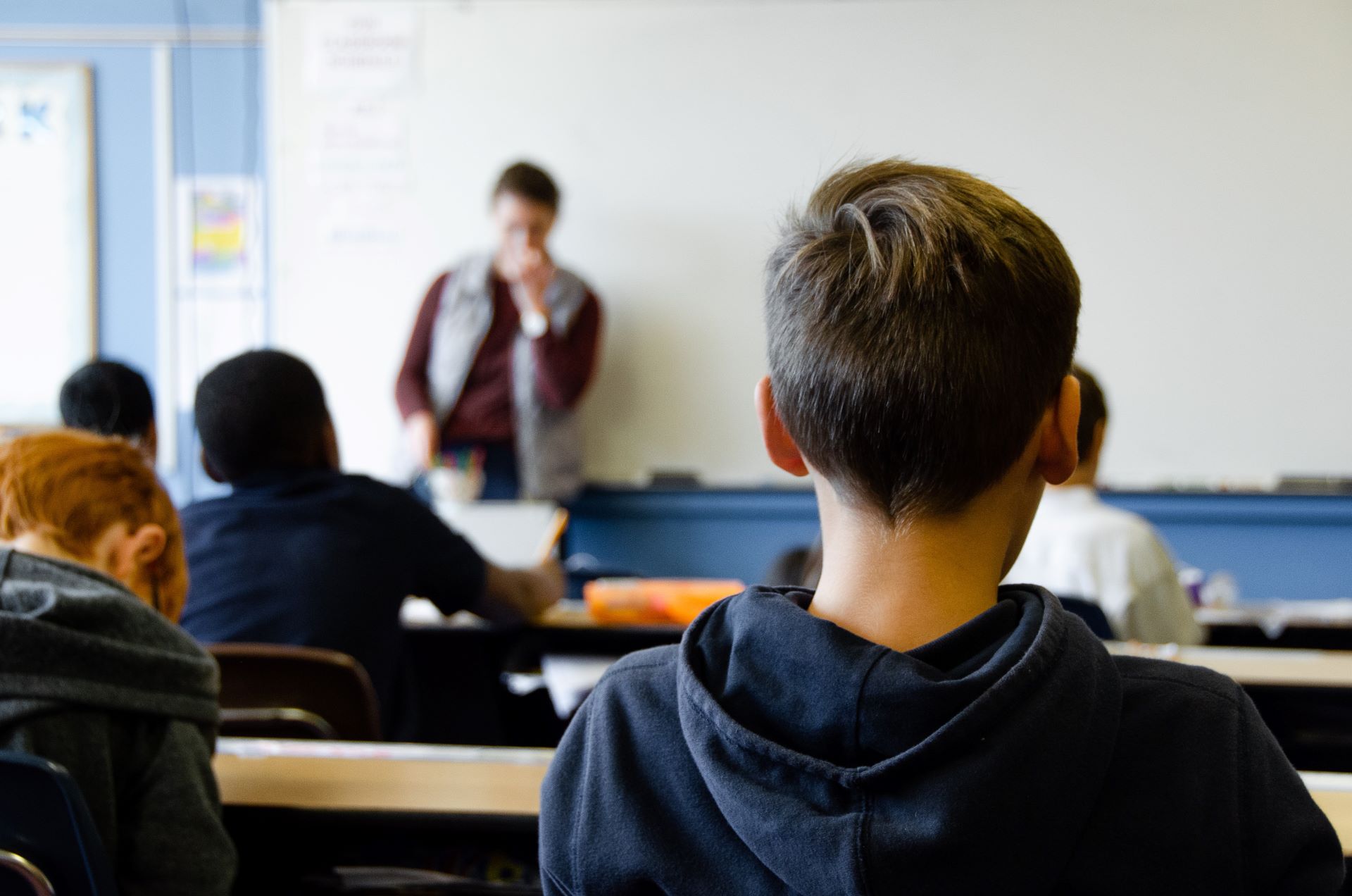 A child sitting at the back of the classroom looking at the teacher standing by a whiteboard.