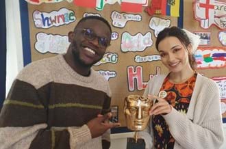De-Graft Mensah and Mirain Iwerydd standing in a classroom smiling while holding a BAFTA trophy.