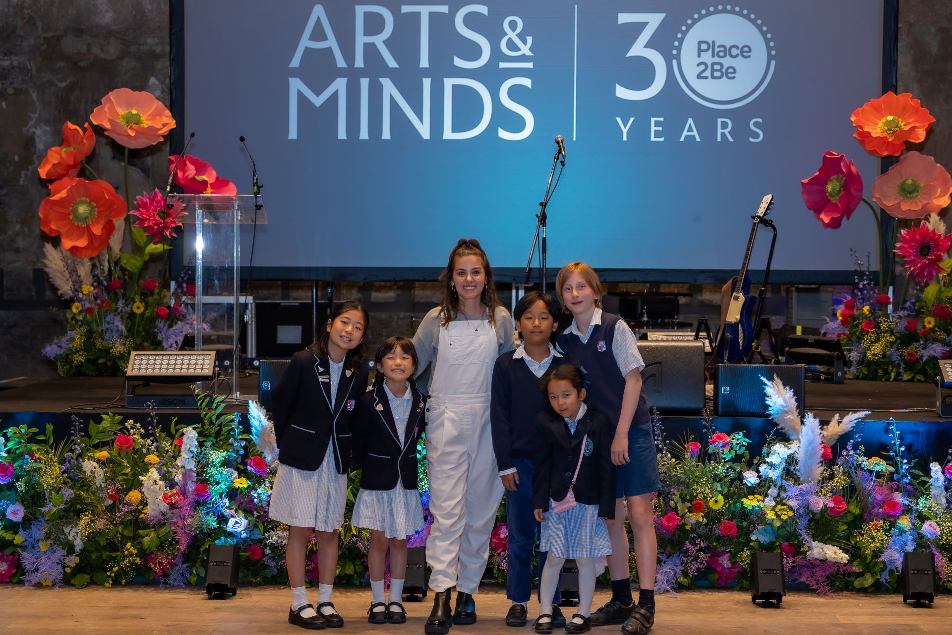 Singer-songwriter Katie Melua standing with a group of pupils from Knightsbridge School in front of the stage at Battersea Arts Centre. Large orange and pink flowers have been placed on either side of the stage.