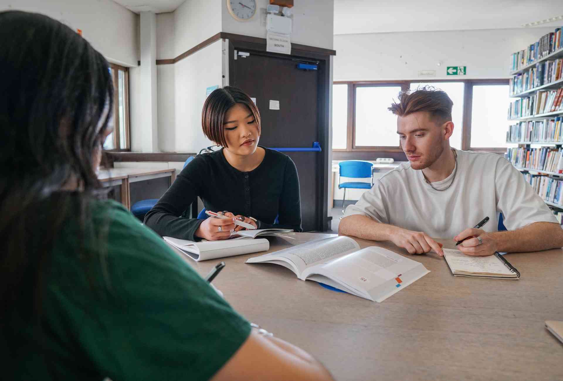 Three university students sitting together looking at a textbook and making notes. They are sitting in a library,