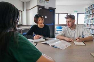 Three university students sitting together looking at a textbook and making notes. They are sitting in a library,