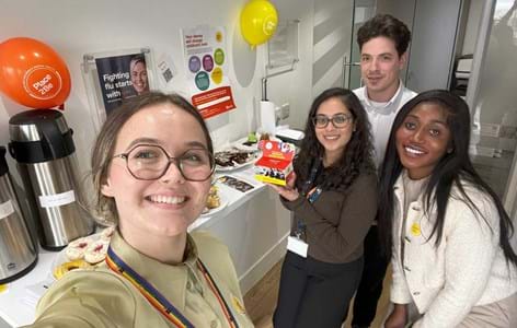 Four young colleagues standing in their workplace having a bake sale.