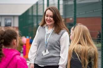 Two young students facing a woman who is smiling at them outside on school grounds. She is wearing a grey vest and a Place2Be lanyard around her neck.