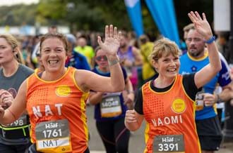 Two women taking part in the Royal Parks Half Marathon. They are wearing Place2Be running vests and waving to people in the crowd.