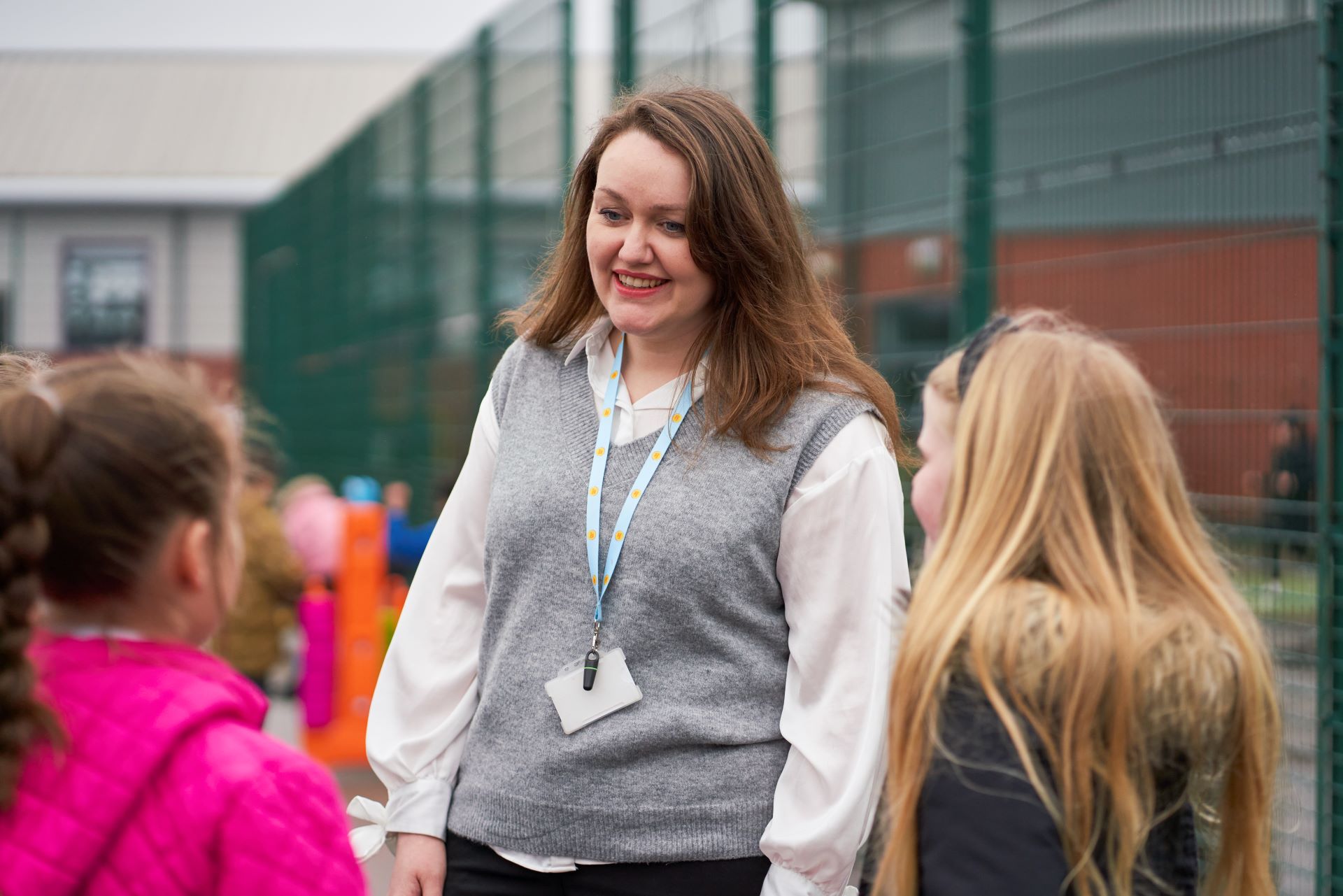 Two young students facing a woman who is smiling at them outside on school grounds. She is wearing a grey vest and a Place2Be lanyard.